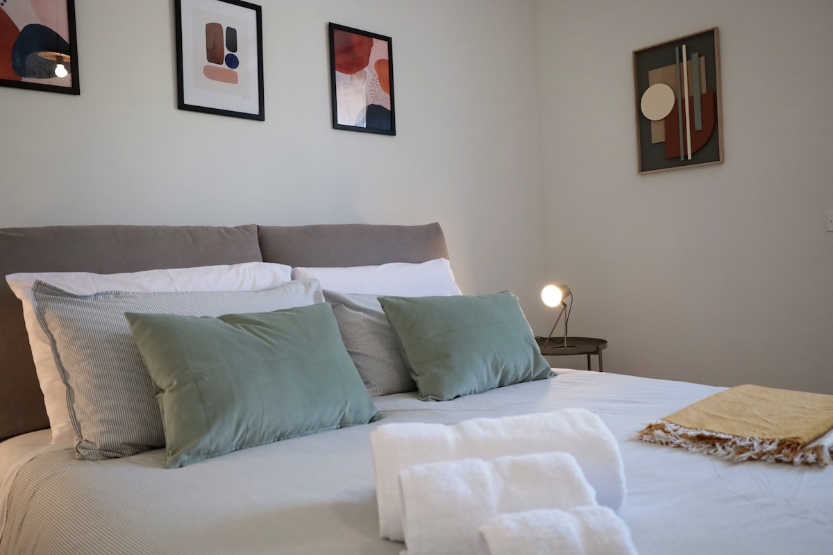 A comfortable sleeping area features a spacious bed with decorative pillows, complemented by art pieces hanging on the walls. Soft towels are neatly arranged at the foot of the bed, and a bedside lamp provides warm lighting, enhancing the room's relaxing ambiance.