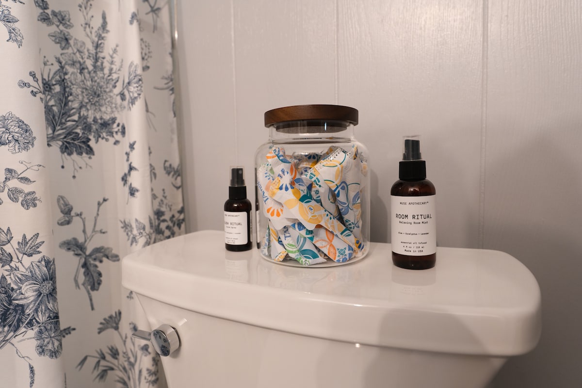 A neatly arranged bathroom countertop features a white toilet with a decorative towel holder. A large glass jar filled with colorful floral-patterned towels sits beside two dark glass spray bottles labeled for room rituals, enhancing the organized and inviting atmosphere.