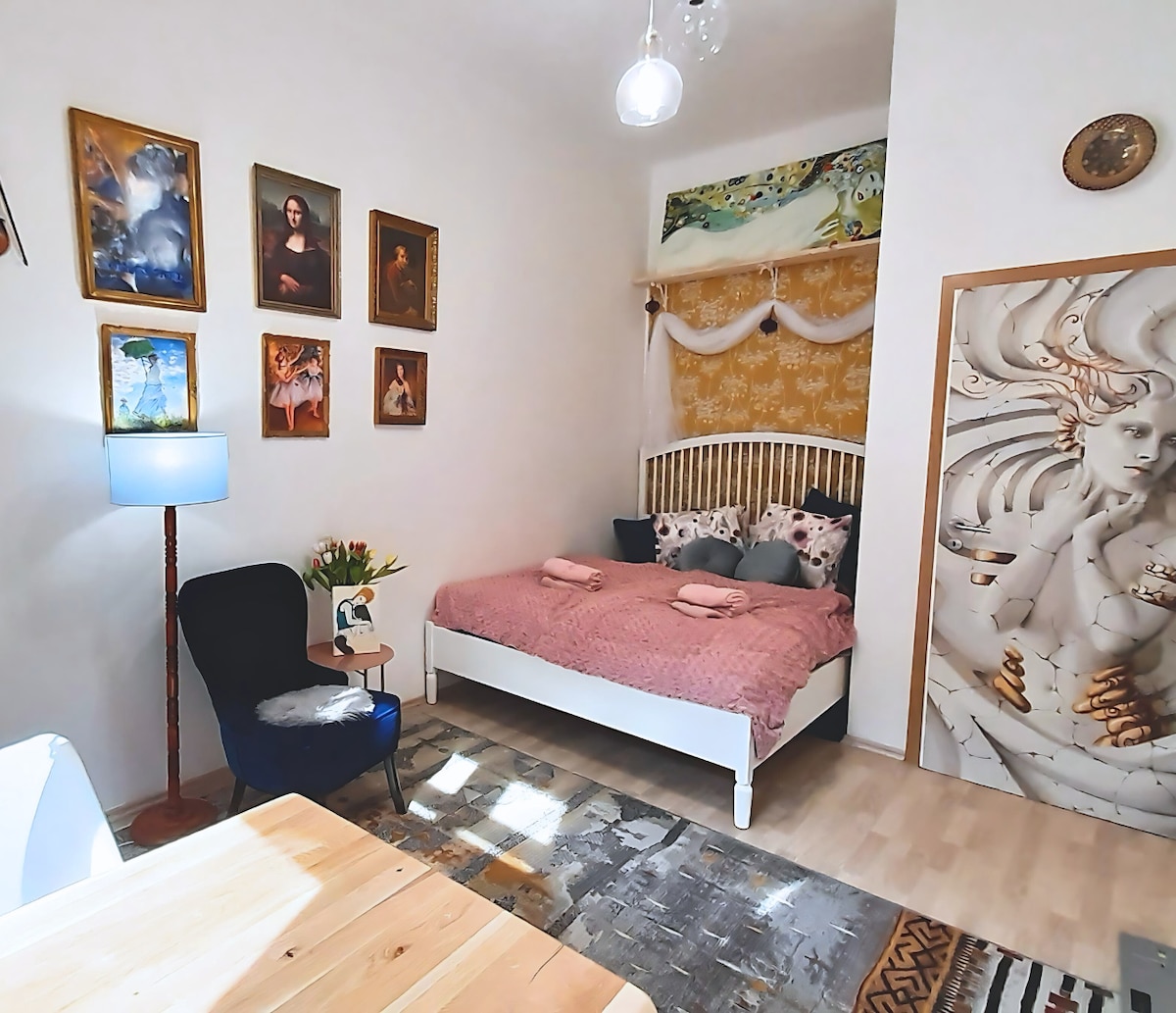 A cozy bedroom is presented, featuring a spacious bed adorned with a pink blanket and decorative pillows. Framed artwork adorns the walls, complemented by a blue floor lamp and a small table with flowers. A chair provides additional seating, enhancing the inviting atmosphere.