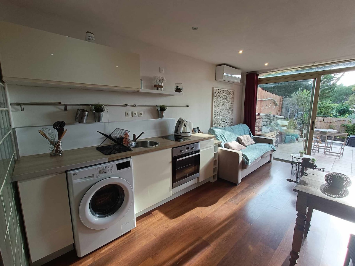 The open kitchen features modern appliances, including a washing machine and stove, complemented by a comfortable sofa. Natural light floods the space through a large sliding door, leading to an outdoor area with garden views. Wood flooring adds warmth to the interior.
