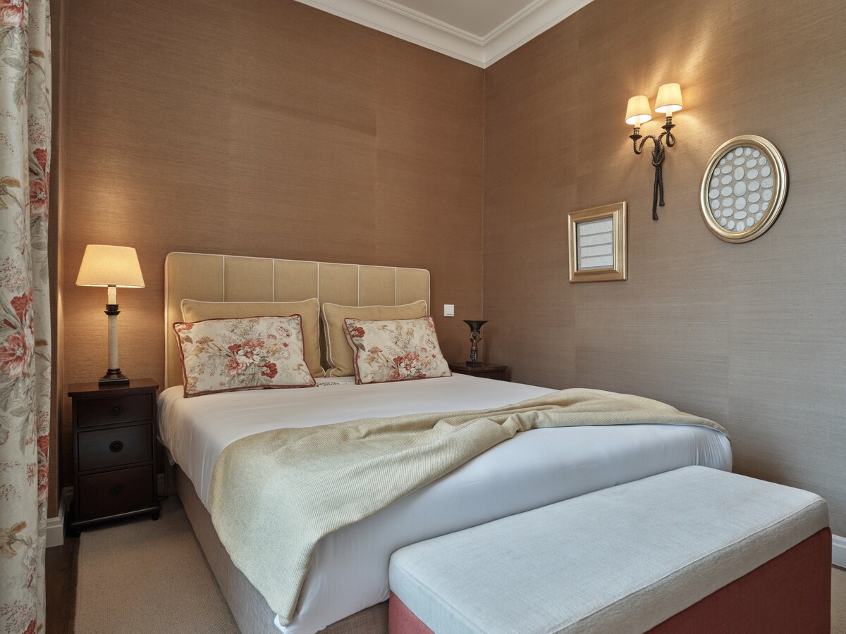 A cozy sleeping area is presented with a spacious bed dressed in neutral linens, flanked by bedside lamps. Decorative pillows with subtle patterns add a touch of elegance. A light-colored bench is placed at the foot of the bed, and soft lighting enhances the warm ambiance.