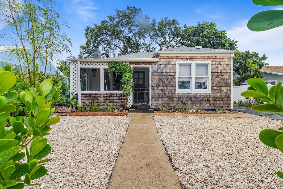 A charming cottage exterior features a pathway leading to the front door, framed by vibrant greenery and a pebbled yard. The textured wood siding adds character, while multiple windows allow natural light to fill the surrounding space.