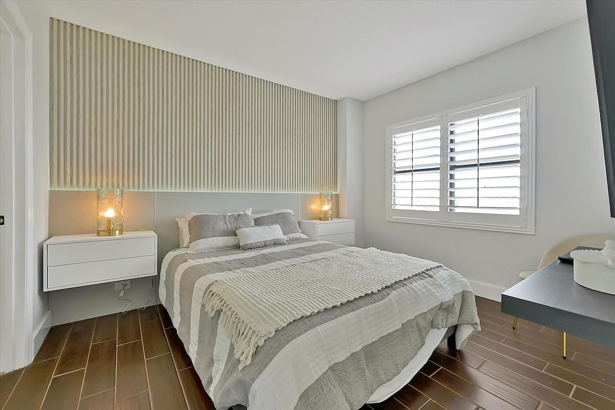 Спалня с двойно легло, дървена табла с подсветка, телевизор на стената, бели нощни шкафчета и самостоятелна баня. Уютен и модерен дизайн.