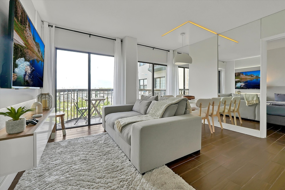 Очарователна всекидневна със светлосив диван, уютно одеяло, пухкав килим и балкон с открит изглед. Отворена концепция с трапезария и кухня.