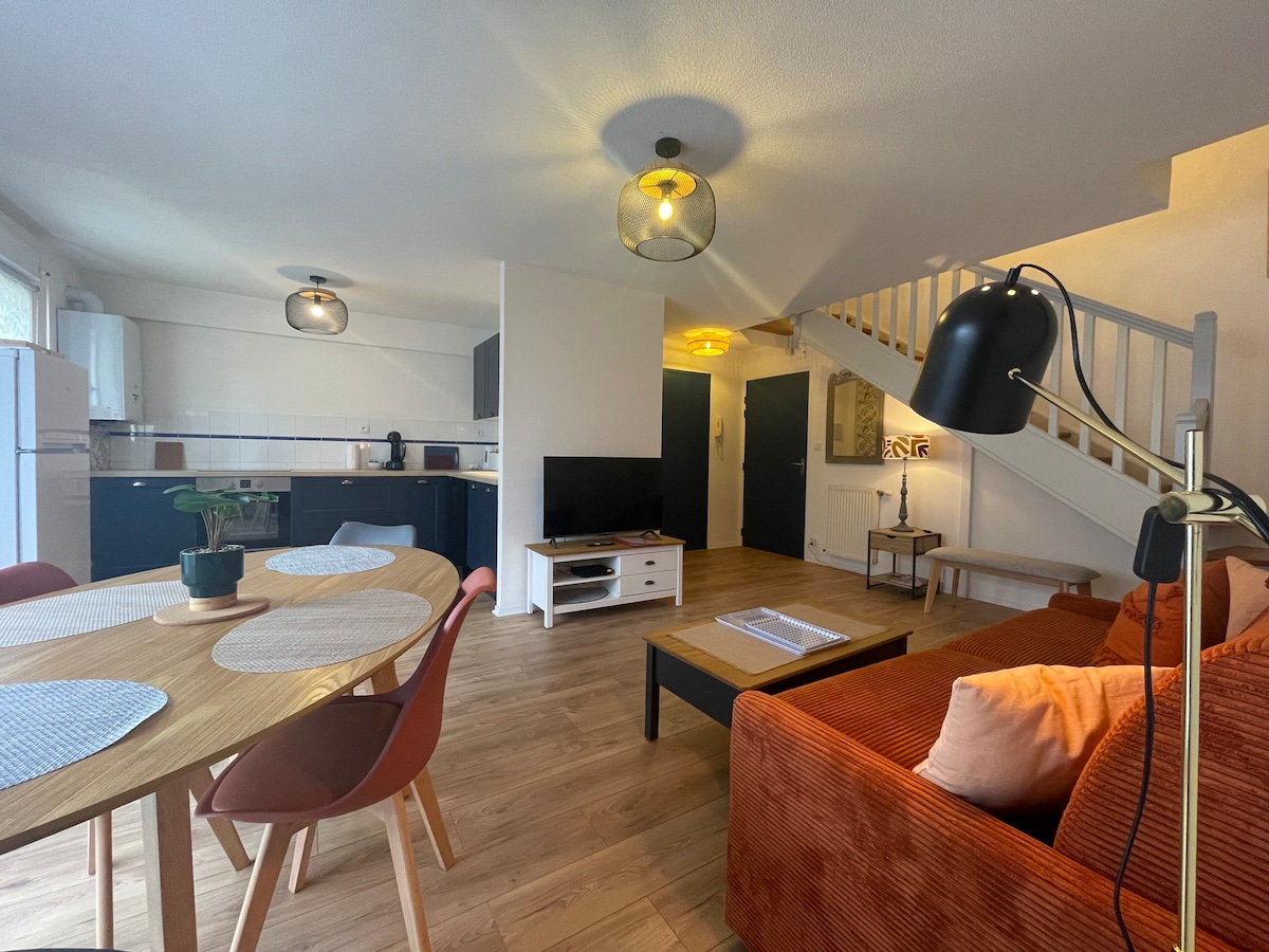 The image features a modern living space with a spacious layout. A round wooden dining table, surrounded by colorful chairs, is positioned near the kitchen. A cozy orange sofa is facing a television, with warm light fixtures enhancing the inviting atmosphere. The area provides access to an upper level.