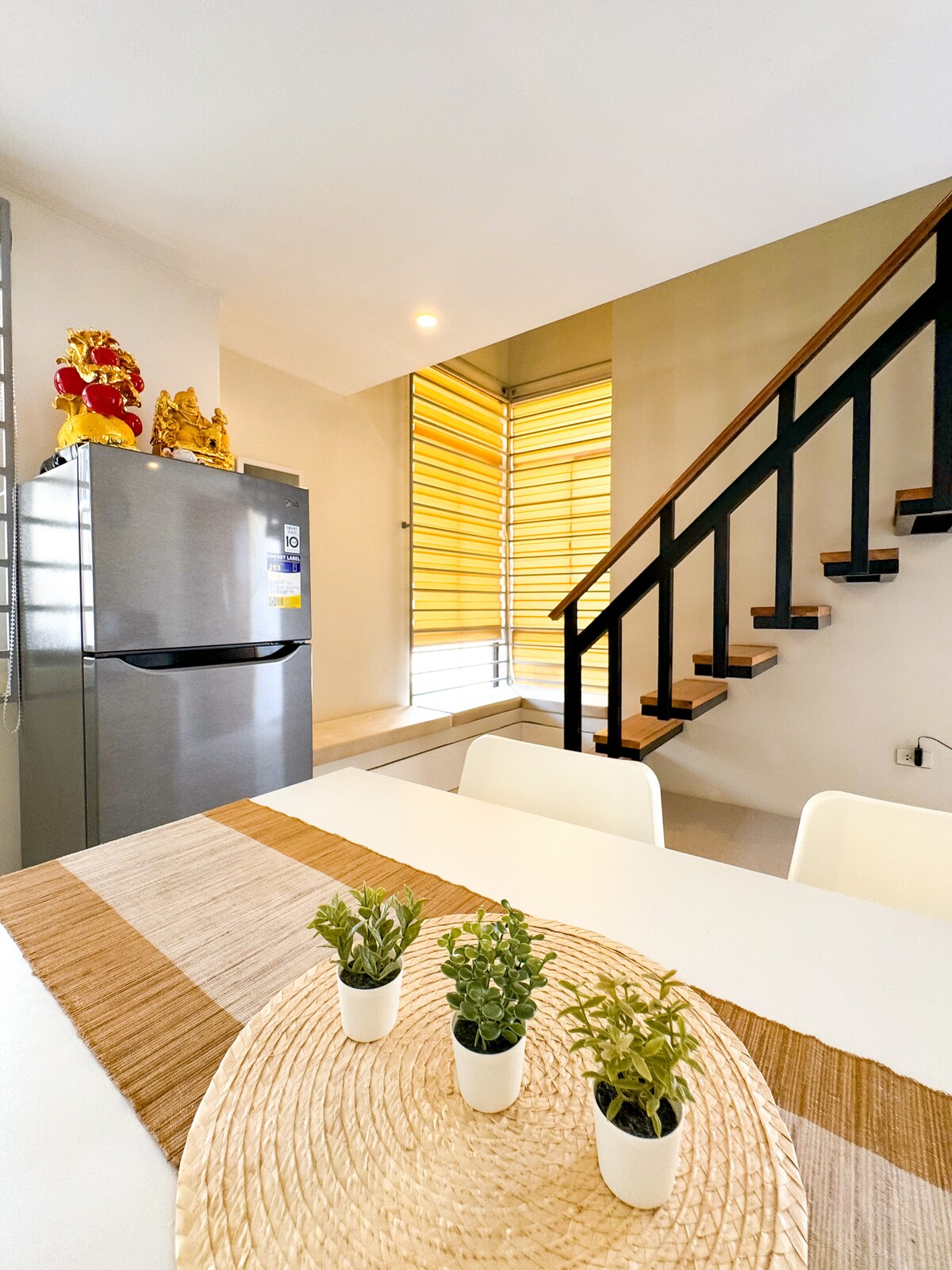 A clean and modern kitchen area features a sleek refrigerator and a dining table adorned with small potted plants. Natural light filters through the window, highlighting the staircase that leads to the upper level.