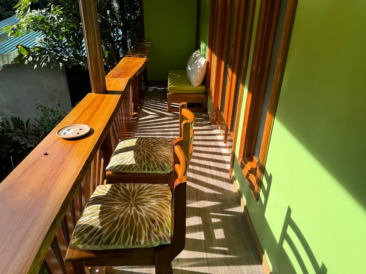 A balcony with wooden furnishings, featuring two chairs with patterned cushions and a small round table. The space is adorned with vibrant green walls, and natural light creates dynamic shadows across the floor, enhancing the connection to the surrounding nature.