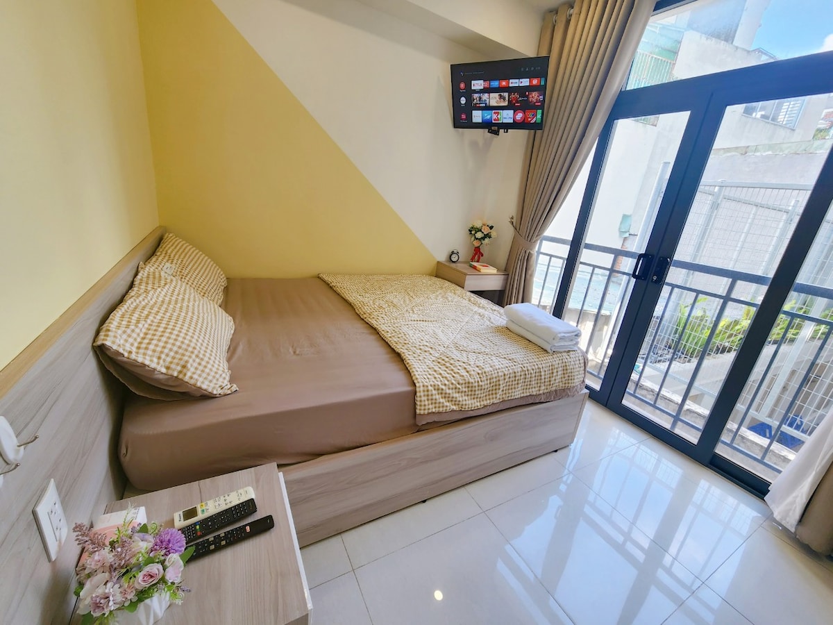 A cozy sleeping area is highlighted by a queen-size bed dressed in neutral bedding. A wall-mounted smart TV is positioned near the bed, and large glass doors provide access to a balcony, allowing natural light to brighten the room. Soft curtains frame the view.