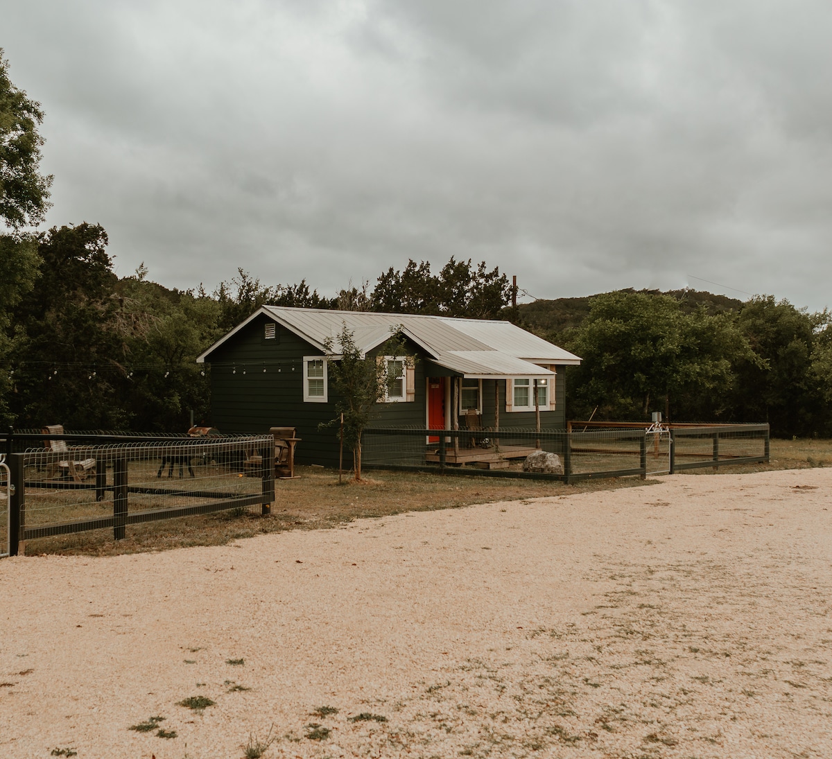 A cozy cabin with green siding and a metal roof is positioned on a spacious lot. The front porch is highlighted by a welcoming red door. A gravel path leads up to the cabin, framed by a black fence and surrounded by lush greenery.