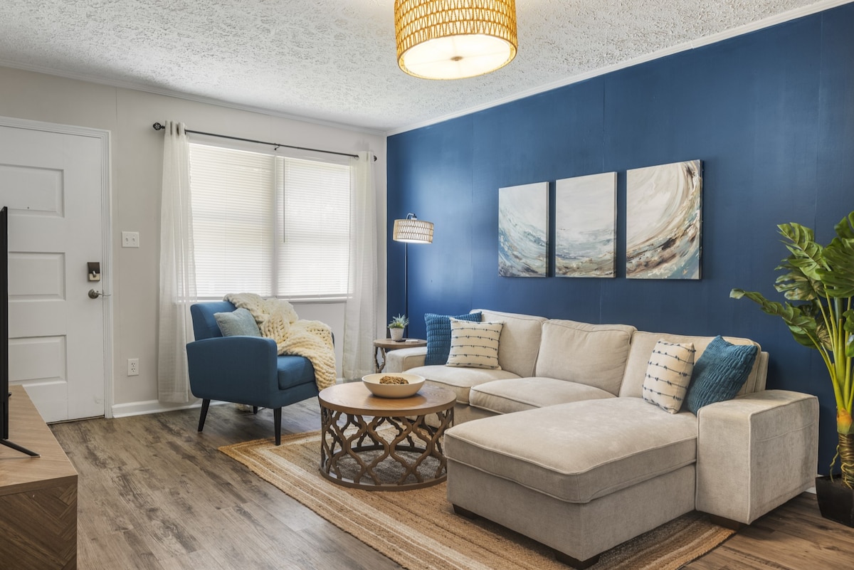 The living room features a blue accent wall complemented by a neutral sectional sofa and a round coffee table. A blue armchair is positioned nearby, with decorative pillows adding a pop of color. Natural light filters through sheer curtains, enhancing the inviting atmosphere.
