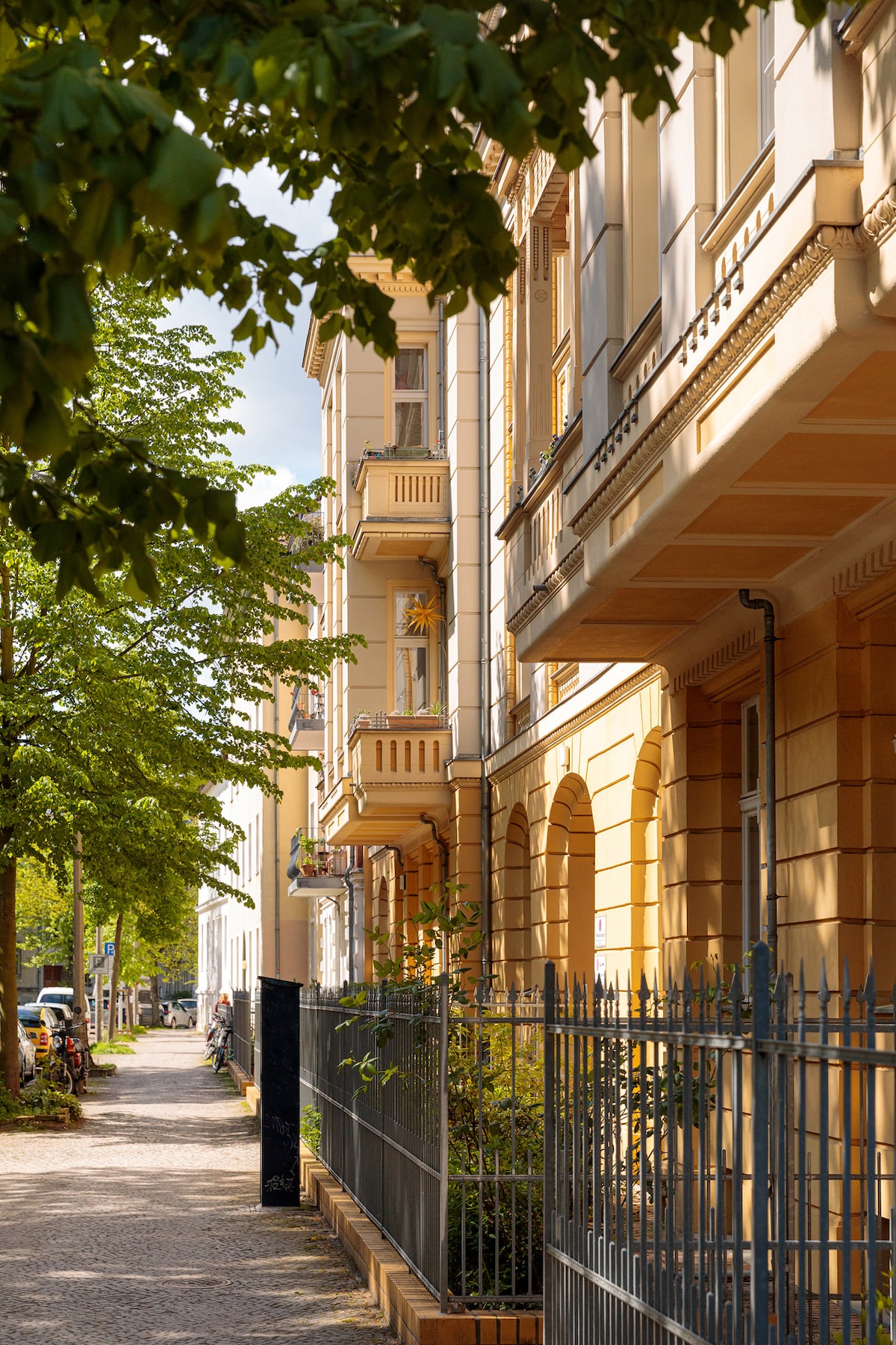 A serene street lined with historic townhouses showcases classic architecture. Lush green trees provide shade along the walkway, while charming details of the buildings, including balconies and arches, are visible. A sense of tranquility is present in this inviting neighborhood.