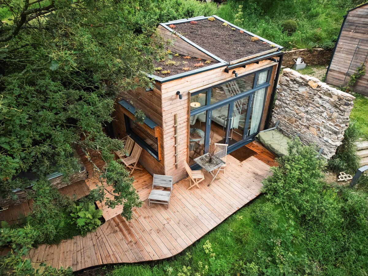 The tiny house features a wooden deck that extends toward the lush greenery, with a mix of outdoor seating options visible. Large glass doors create an open feel, allowing natural light to penetrate the interior space. The green roof integrates the structure into its natural surroundings.