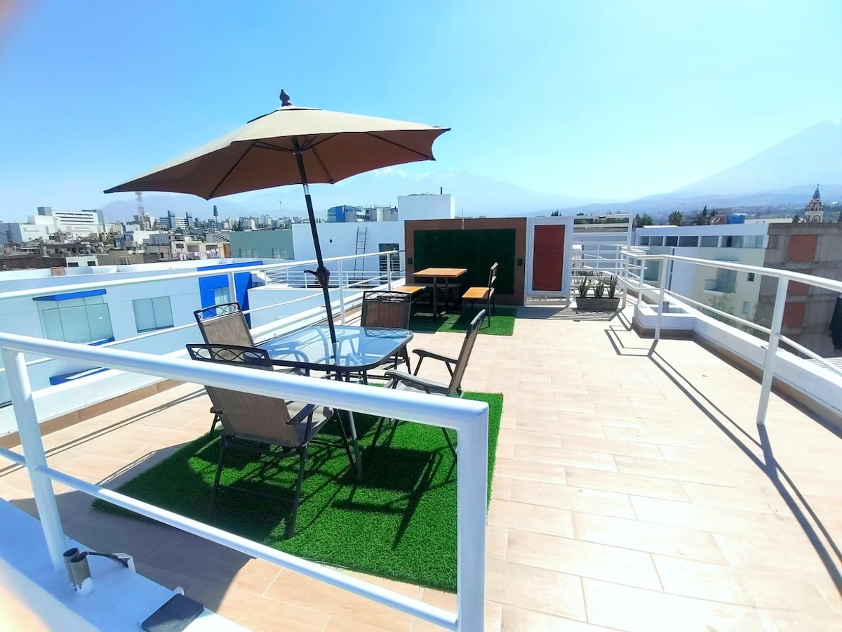 A rooftop terrace is shown, featuring a set of outdoor chairs and a table under a large umbrella. The area is surrounded by greenery, with a view of the skyline and mountains in the background, providing a relaxing space for guests.