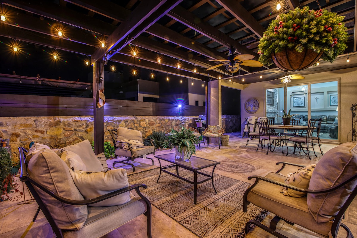 An outdoor living space features comfortable seating arranged around a central table, illuminated by twinkling string lights. A ceiling fan hangs above, while potted plants and decorative elements add a touch of greenery to the setting.