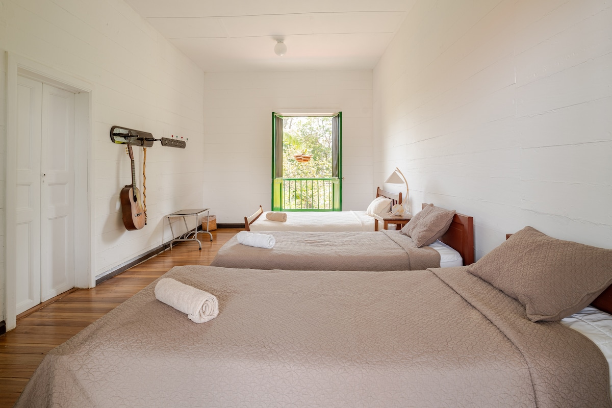 A spacious room is presented with two large beds, each featuring neutral bedding. Light enters through an open window adorned with green shutters, offering views of nature. Wooden floors complement the light-colored walls. A guitar is hung on the wall, and towels are neatly rolled by the beds.