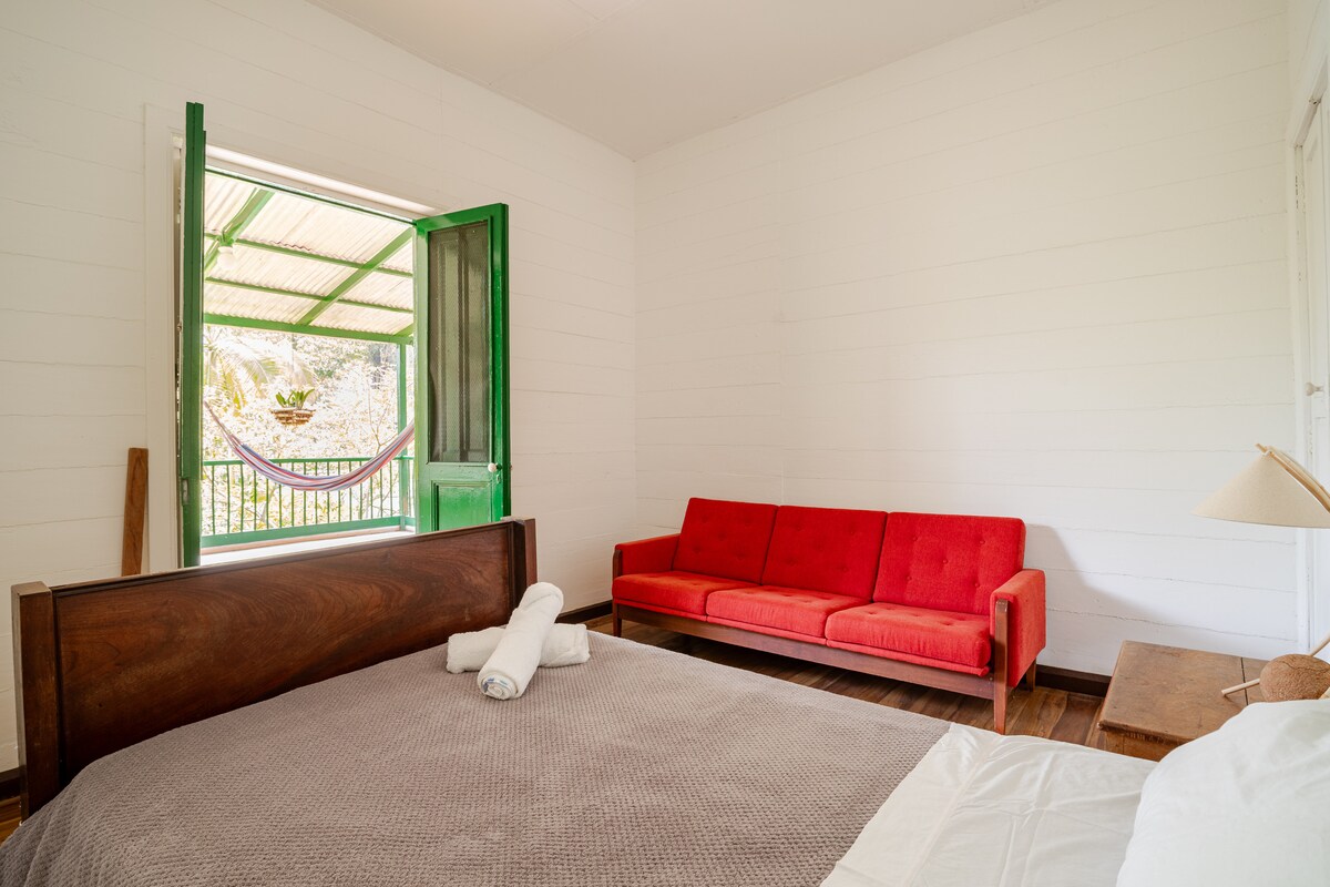 A spacious bedroom features a large bed with neutral-colored linens. A bright red sofa adds a pop of color. An open green door leads to a covered terrace, inviting natural light and fresh air into the room.