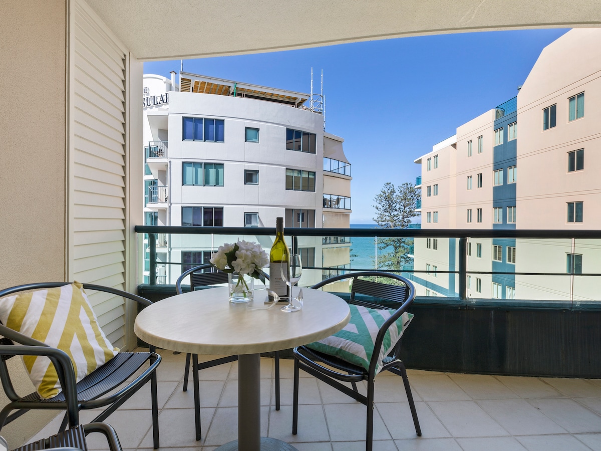 A balcony features a round table and four chairs, two adorned with cushions. A bottle of wine sits on the table alongside two glasses. The background showcases a clear blue sky and views of surrounding buildings and trees, providing a relaxed outdoor setting.