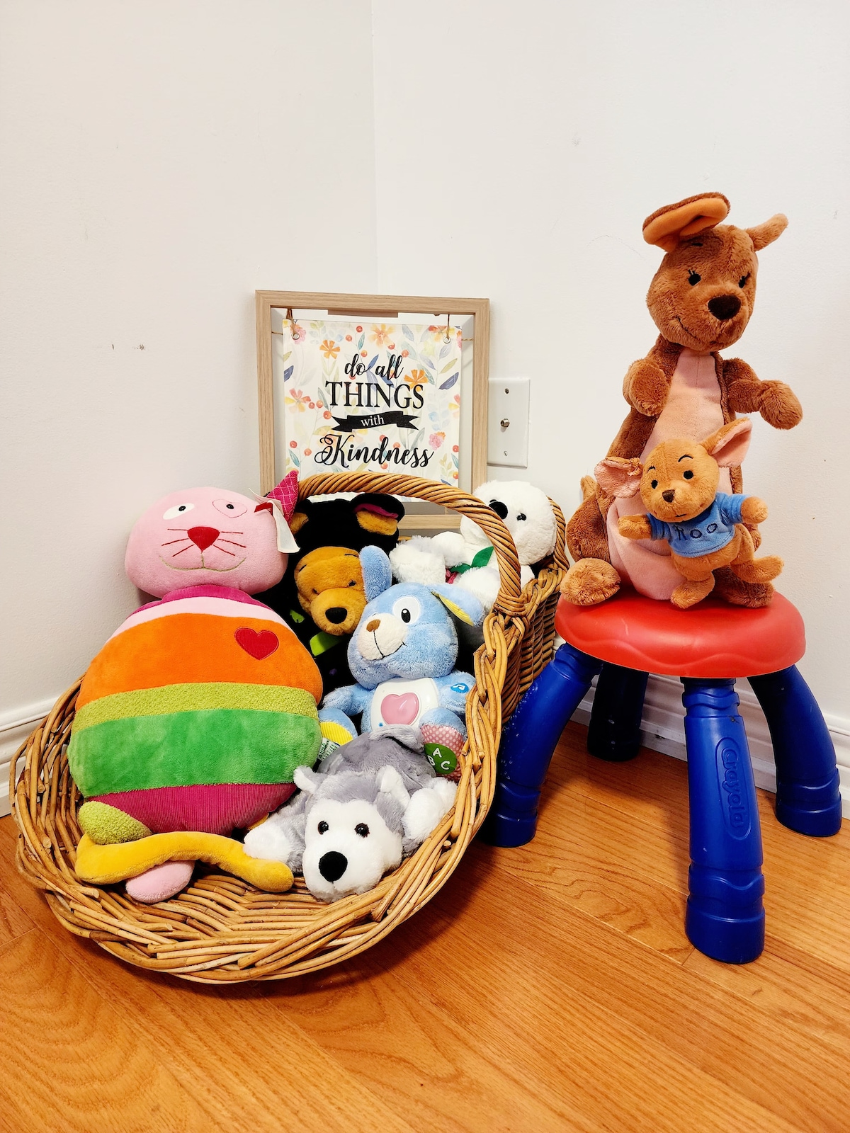 A colorful arrangement of stuffed toys is placed in a woven basket, featuring plush animals in various sizes and hues. A framed sign with the text 'Do All Things With Kindness' is positioned above the basket, accompanied by a small red stool supporting a plush dog toy.