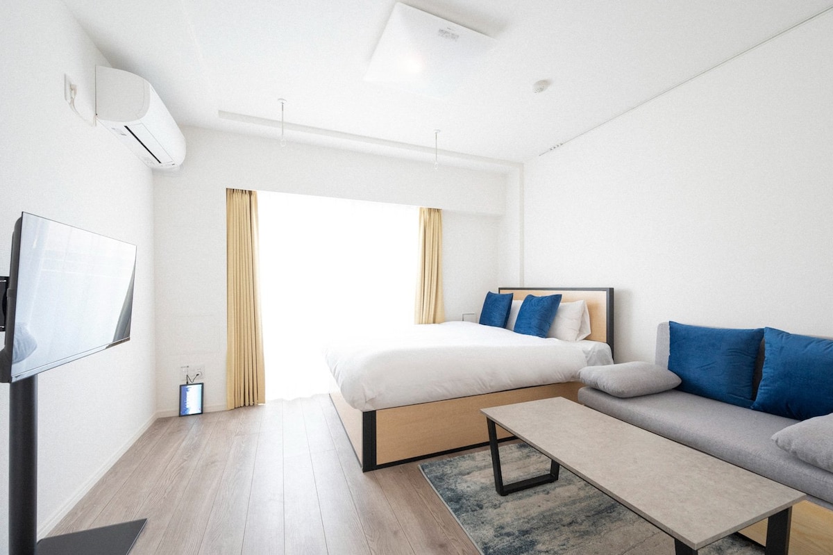 A bright room features a spacious bed with white linens and decorative blue cushions. A gray coffee table is positioned in front, and a television stands nearby. Large windows with light curtains allow natural light to fill the space, enhancing the minimalist design.