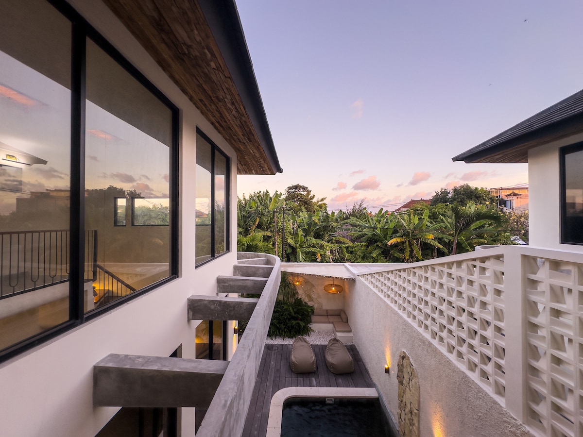 A modern two-story villa showcases a private outdoor space with a serene swimming pool. Soft lounge chairs are positioned beside the water, surrounded by lush tropical vegetation. Large windows reflect the evening sky, enhancing the tranquil ambiance of the retreat.
