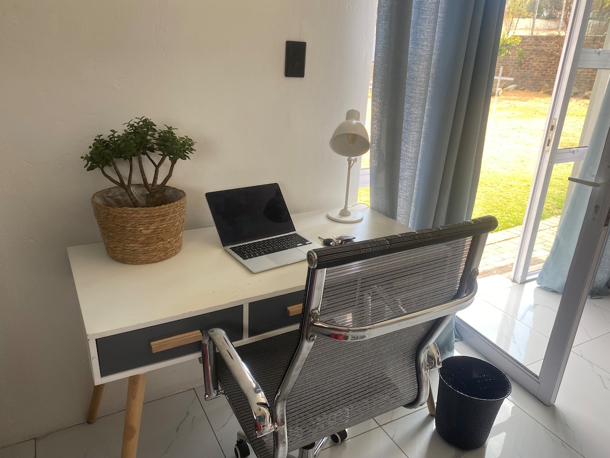 A workspace is presented featuring a sleek desk with a minimalist design. A laptop rests on the surface alongside a contemporary lamp and a potted plant. Natural light enters through a nearby glass door, illuminating the clean, tiled floor and view of the garden outside.
