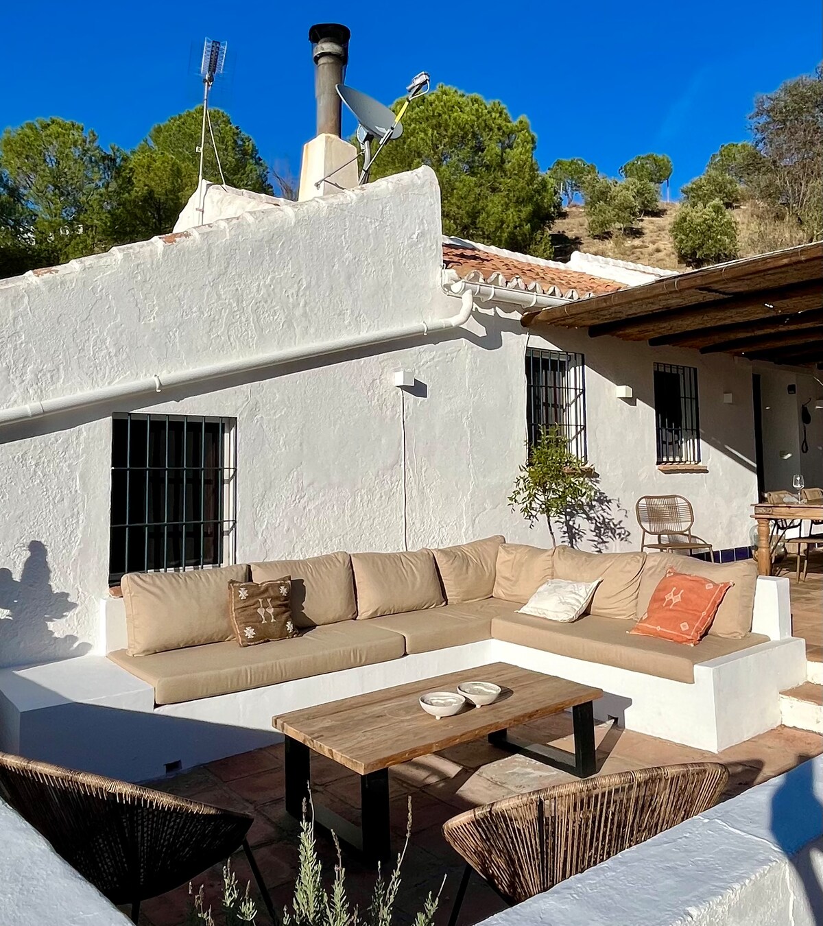 A comfortable outdoor lounge area features a large sectional sofa with soft cushions, ideal for relaxation. In the background, an inviting dining setup is visible, complemented by greenery and the surrounding landscape. The structure's white walls reflect the sunlight, enhancing the tranquil atmosphere.