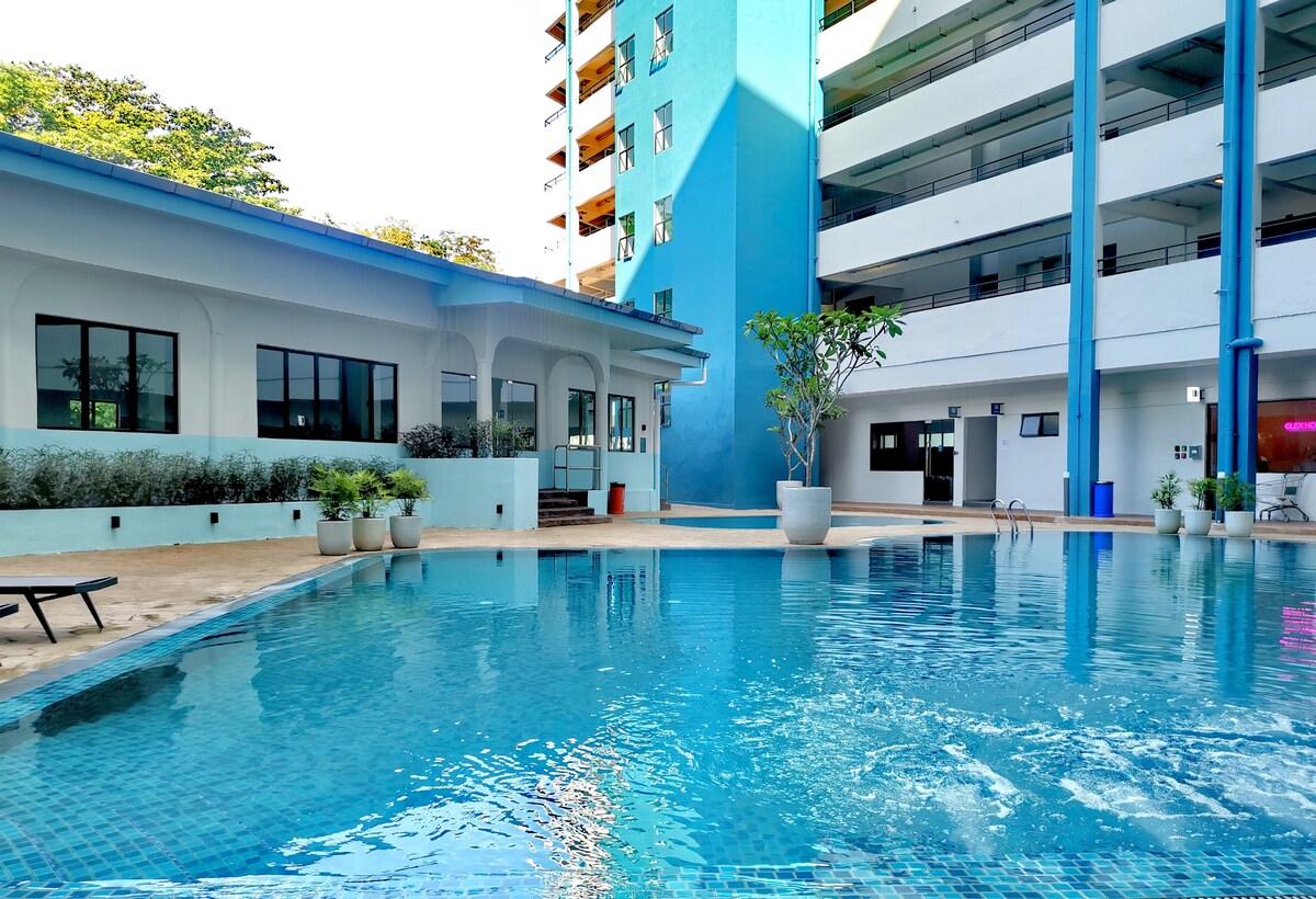 A clear swimming pool reflects the sun, surrounded by neatly arranged lounge chairs and potted plants. The blue building in the background features large windows, enhancing the inviting atmosphere of the shared outdoor space.