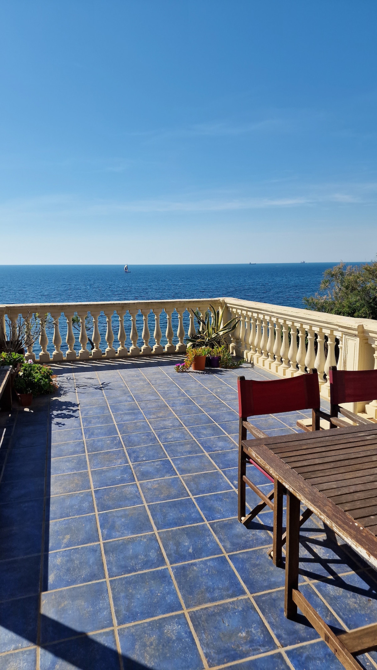 A spacious private terrace overlooks the sea, featuring tiled flooring and a wooden dining set. Potted plants add a touch of greenery, while the expansive view showcases the horizon with boats in the distance under a clear blue sky.