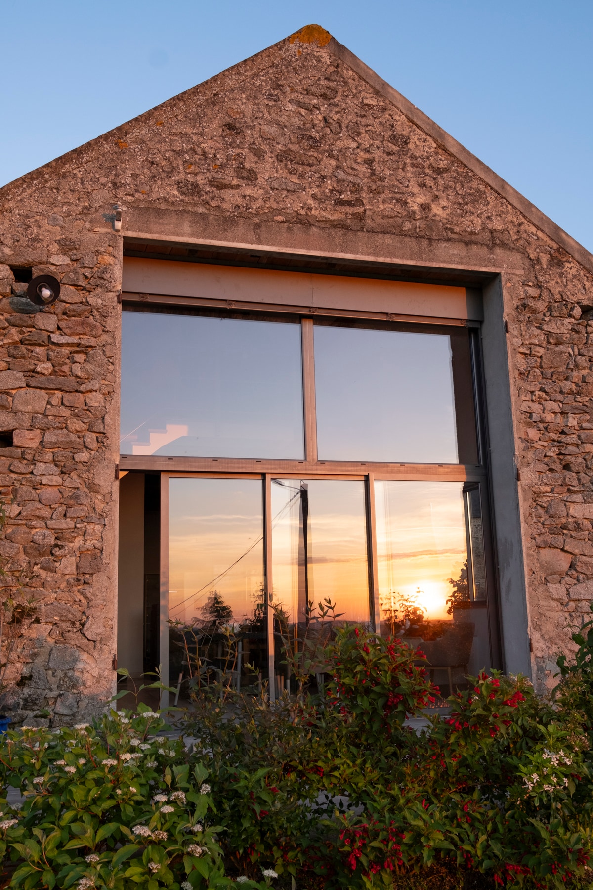 The exterior of the stone building features a large glass facade reflecting a vibrant sunset. Surrounding the structure, lush greenery and colorful flowers contribute to the serene environment, while the silhouette of trees is visible in the background.