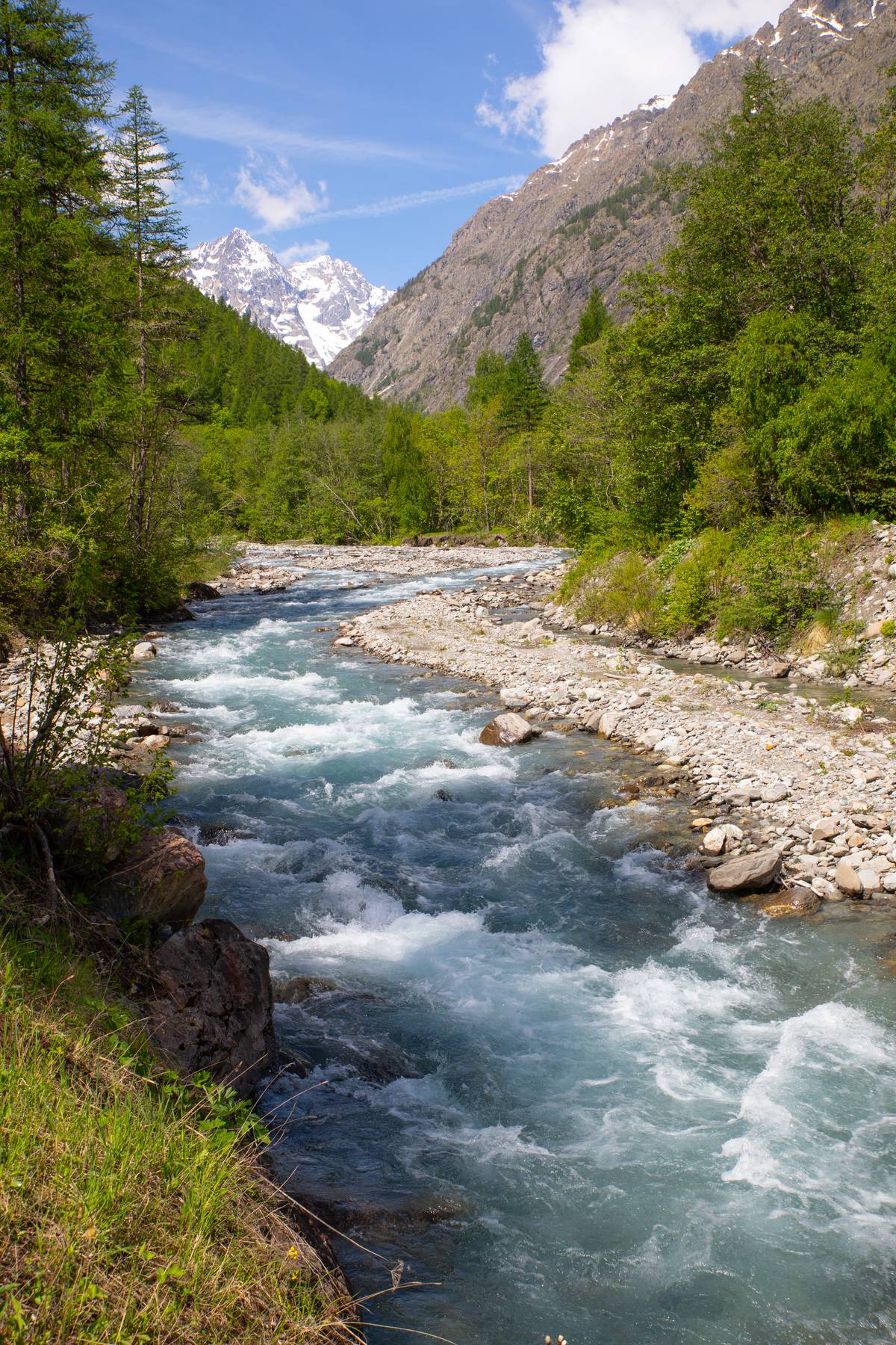 A flowing river winds through a mountainous landscape, flanked by lush greenery and rocky banks. Snow-capped peaks rise in the background against a clear blue sky, creating a serene natural setting that invites exploration.