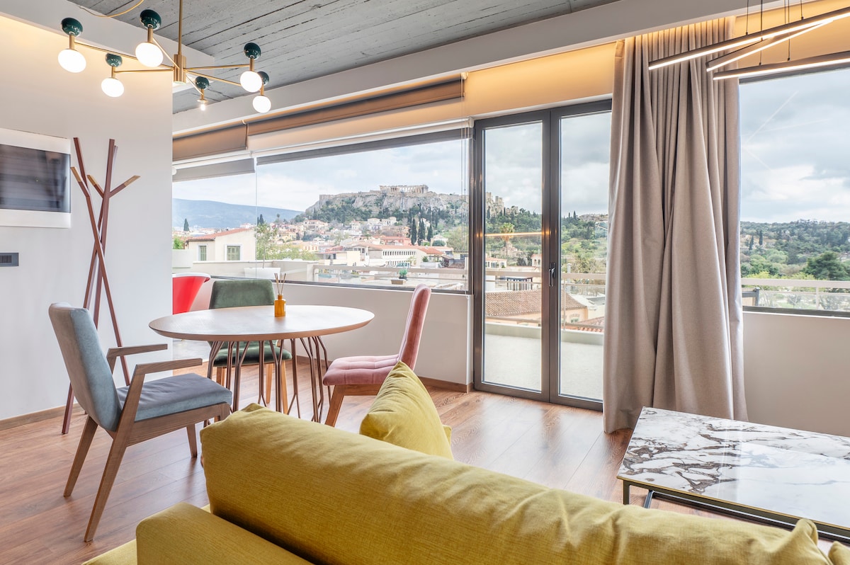 A spacious living area features large windows offering views of the Acropolis. A round dining table is surrounded by colorful chairs, while a comfortable sofa and a sleek coffee table enhance the open atmosphere. Natural light floods the space, creating a bright ambiance.