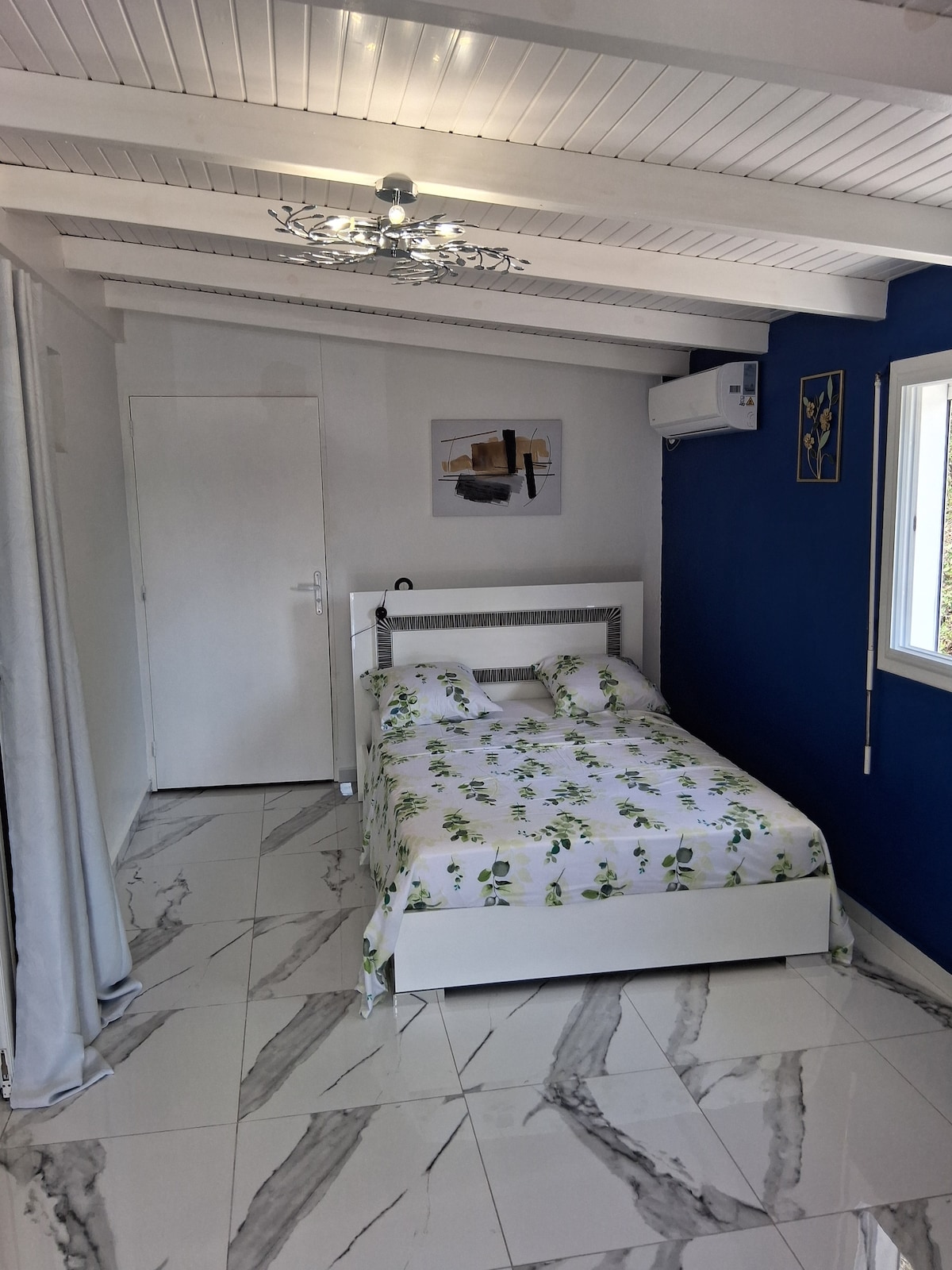 A well-lit bedroom features a white bed dressed in floral linens. A modern ceiling fixture provides illumination, while a blue accent wall adds a pop of color. A closet door and a large window are visible, allowing natural light to enhance the space.