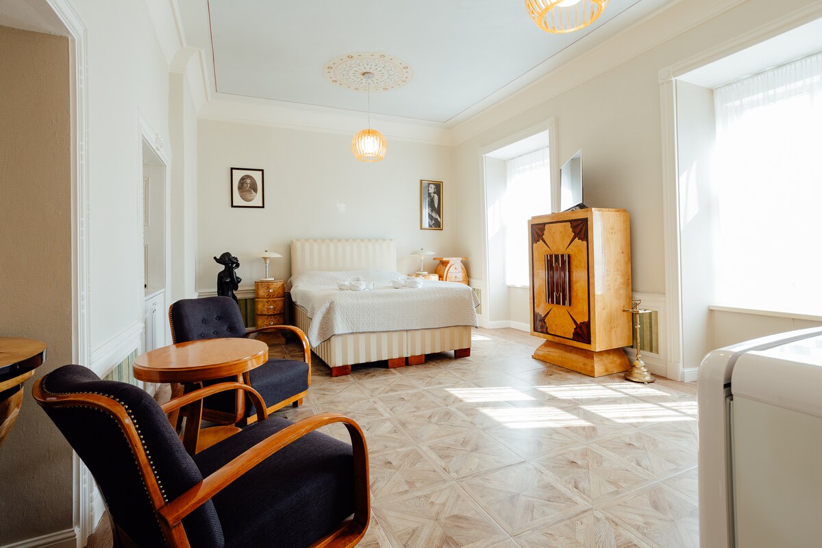 A spacious room features a large bed dressed in neutral linens and a stylish wooden cabinet. Two armchairs are positioned beside a small table, and a TV stands on a contemporary piece of furniture. Natural light filters through two windows adorned with sheer curtains.