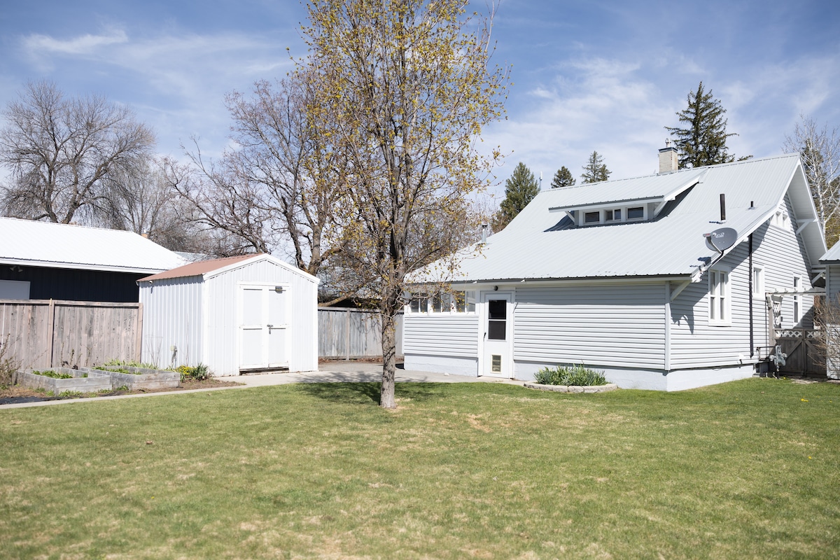 A well-maintained backyard is depicted, featuring a grassy area bordered by a wooden fence. A small shed is visible, and a tree adds to the space's serenity. Sunlight illuminates the exterior of the house, highlighting its light-colored siding and metal roof.