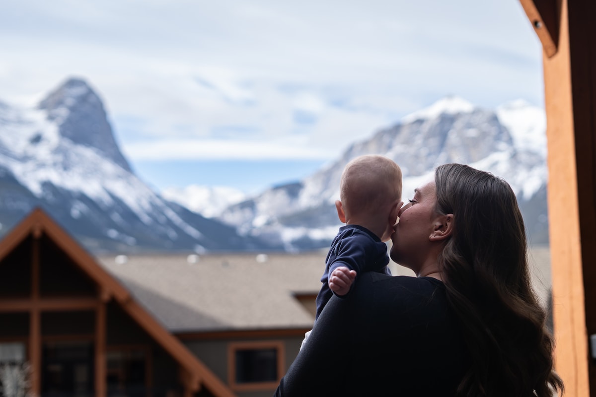 A person is seen holding a baby, with majestic mountains in the background. The peaks are capped with snow, and the sky displays a mix of clouds and blue. The building's wooden structure is partially visible, contributing to a serene environment.