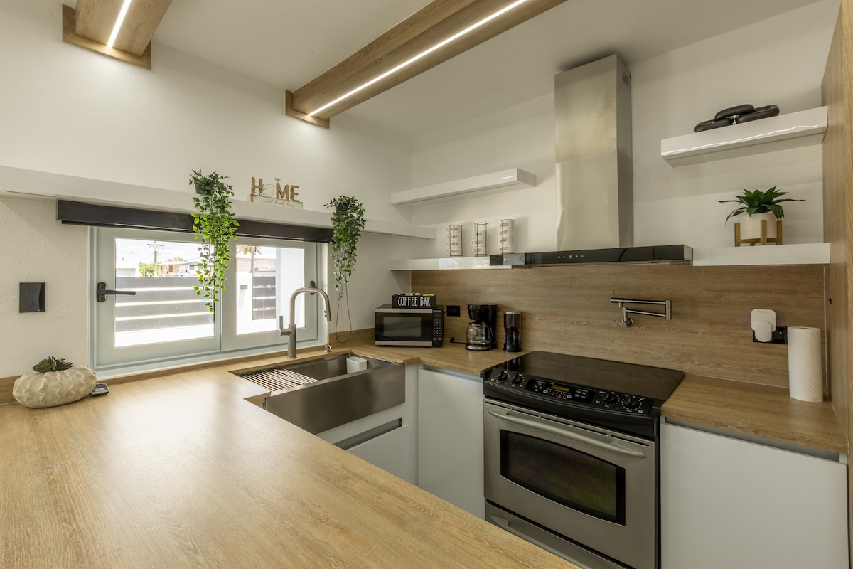 A modern kitchen features sleek cabinetry and a countertop made of light wood. Stainless steel appliances, including an oven and microwave, are complemented by minimalist shelving. Greenery is added with potted plants near the window, which allows natural light to fill the space.