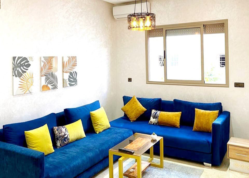 A living area features a vibrant blue sectional sofa complemented by yellow cushions. A wooden coffee table sits in front, enhancing the space. Three framed artworks adorn the wall, and a large window allows natural light to fill the room.