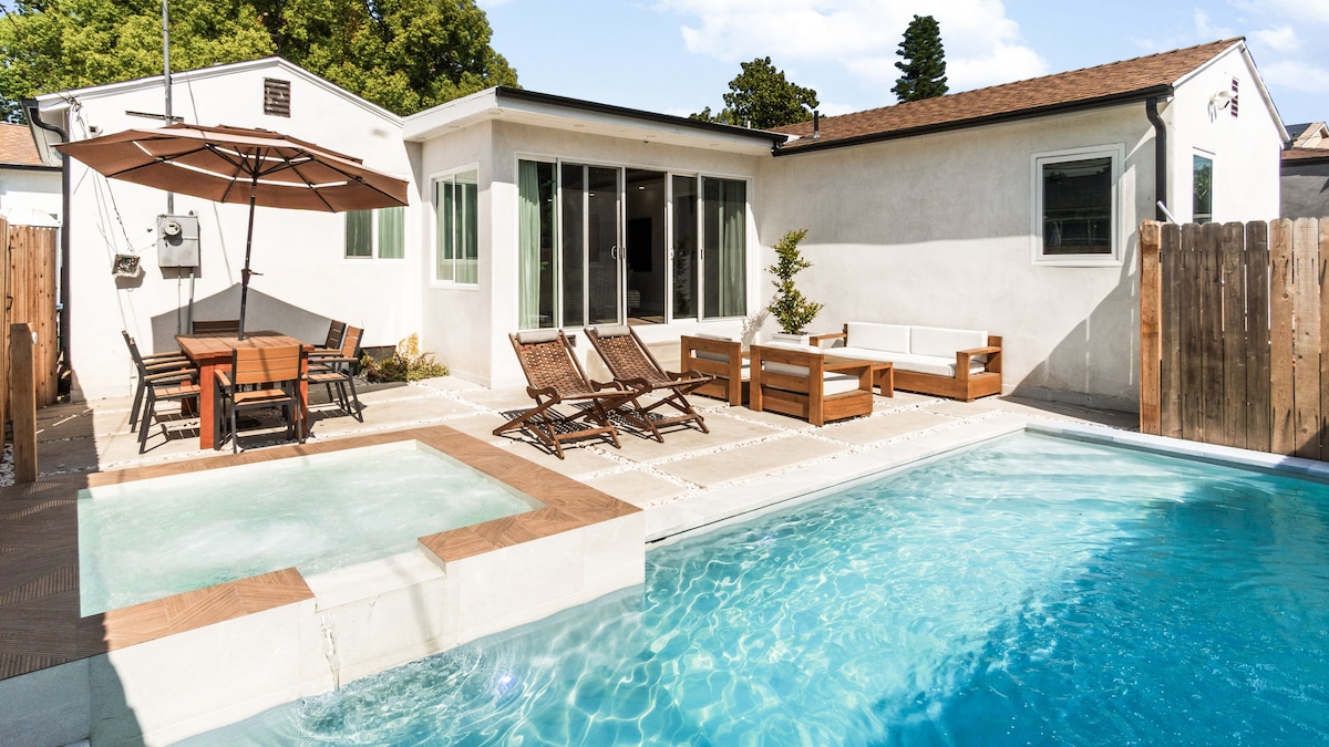 A spacious backyard area features a refreshing pool and a connected spa, surrounded by a stone patio. Poolside seating includes lounge chairs and a wooden dining table with umbrella. The adjacent house is visible, showcasing large windows that invite natural light.