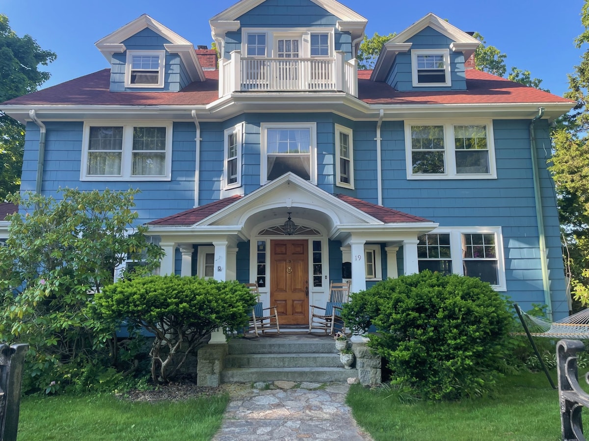 A charming three-story house is framed by lush greenery and well-maintained shrubs. The entrance features a spacious stone pathway leading to double doors, adorned by a decorative overhang. A small porch area includes inviting seating, creating a welcoming first impression.