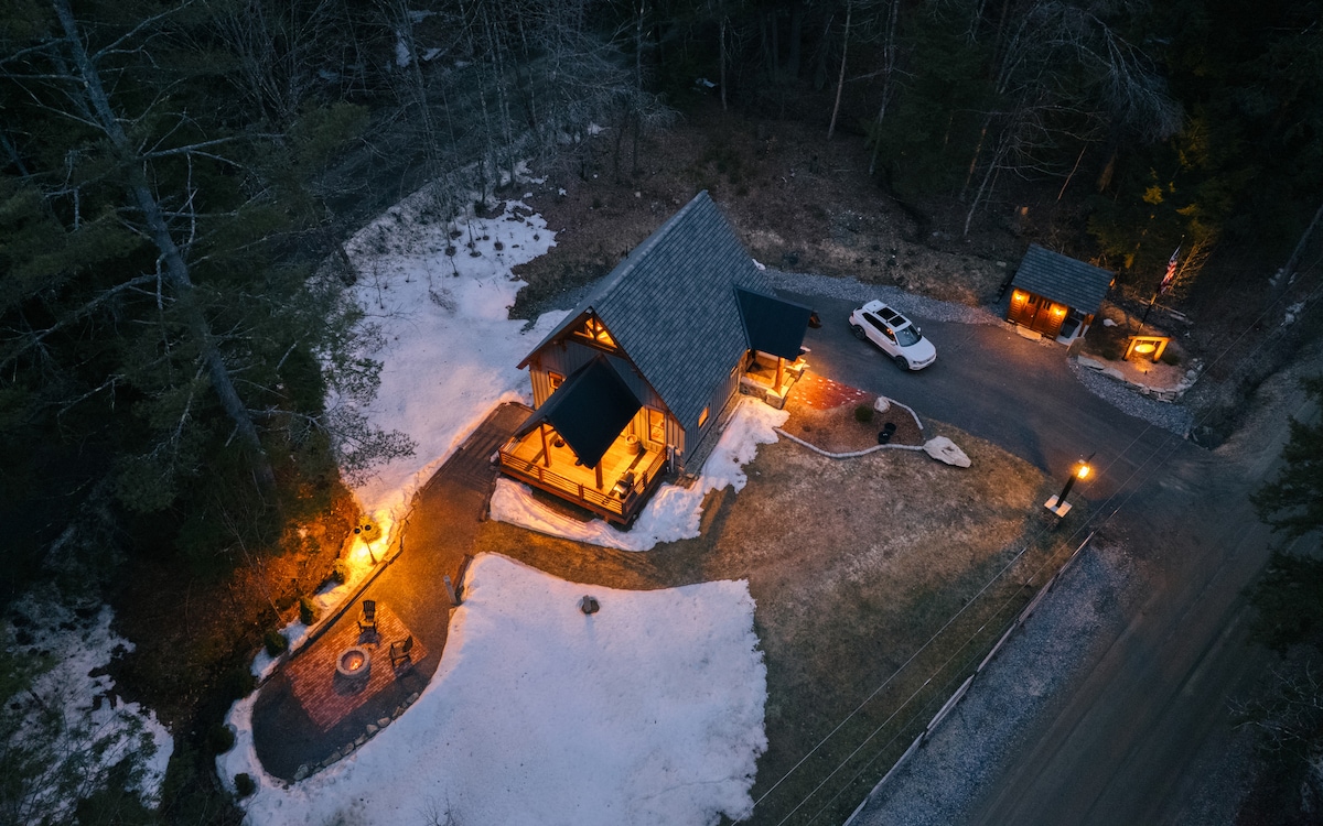An aerial view captures the cabin nestled in a snowy landscape, showcasing warm lighting from exterior fixtures. The cabin features a stone patio with a fire pit and seating arrangements, while nearby vehicles are parked, highlighting the accessibility of the property.