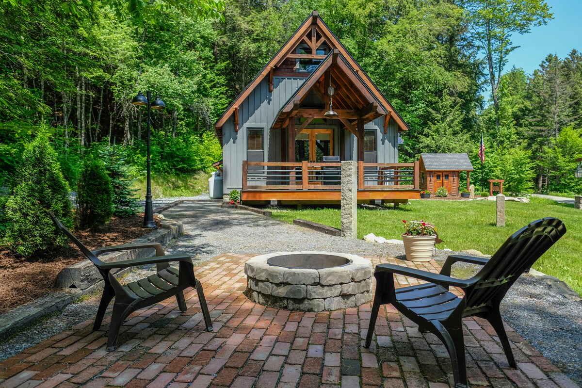 A charming cabin with a wooden facade is framed by lush greenery. Two black lounge chairs are positioned around a round stone fire pit, placed on a patterned brick patio. A well-kept lawn extends in front, adorned with a flower pot and a small outbuilding.