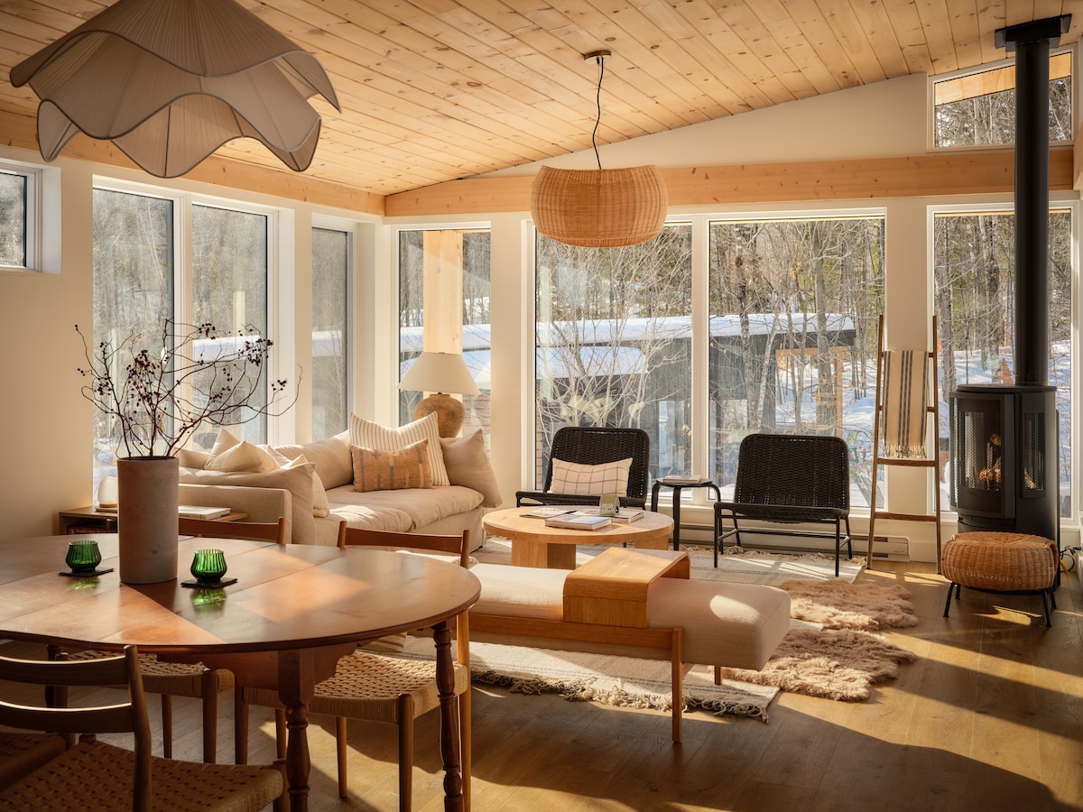 A light-filled living area showcases large windows that invite natural light and views of the surrounding nature. Modern furnishings, including a cozy sofa, a wooden coffee table, and a Scandinavian-style gas fireplace, create a welcoming space. A dining table with wicker chairs is positioned nearby.