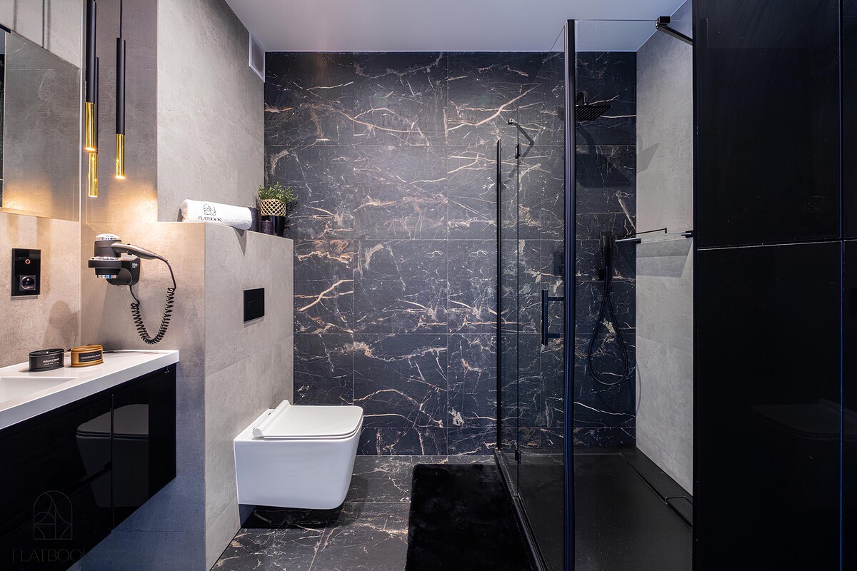 A modern bathroom features elegant dark marble walls with gold veining. A glass shower enclosure and a freestanding white bathtub are present. Minimalist black cabinetry and stylish lighting accents complete the refined design, providing a sleek and sophisticated appearance.