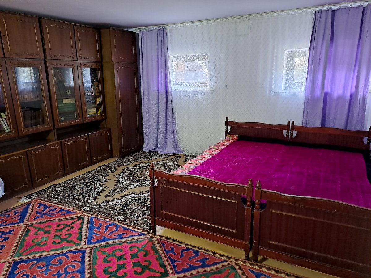 The spacious room features a large bed with a vibrant purple coverlet and decorative pillows. A dark wooden wardrobe with glass doors stands against one wall, complemented by a colorful patterned rug that adds warmth to the space. Natural light filters through sheer curtains.