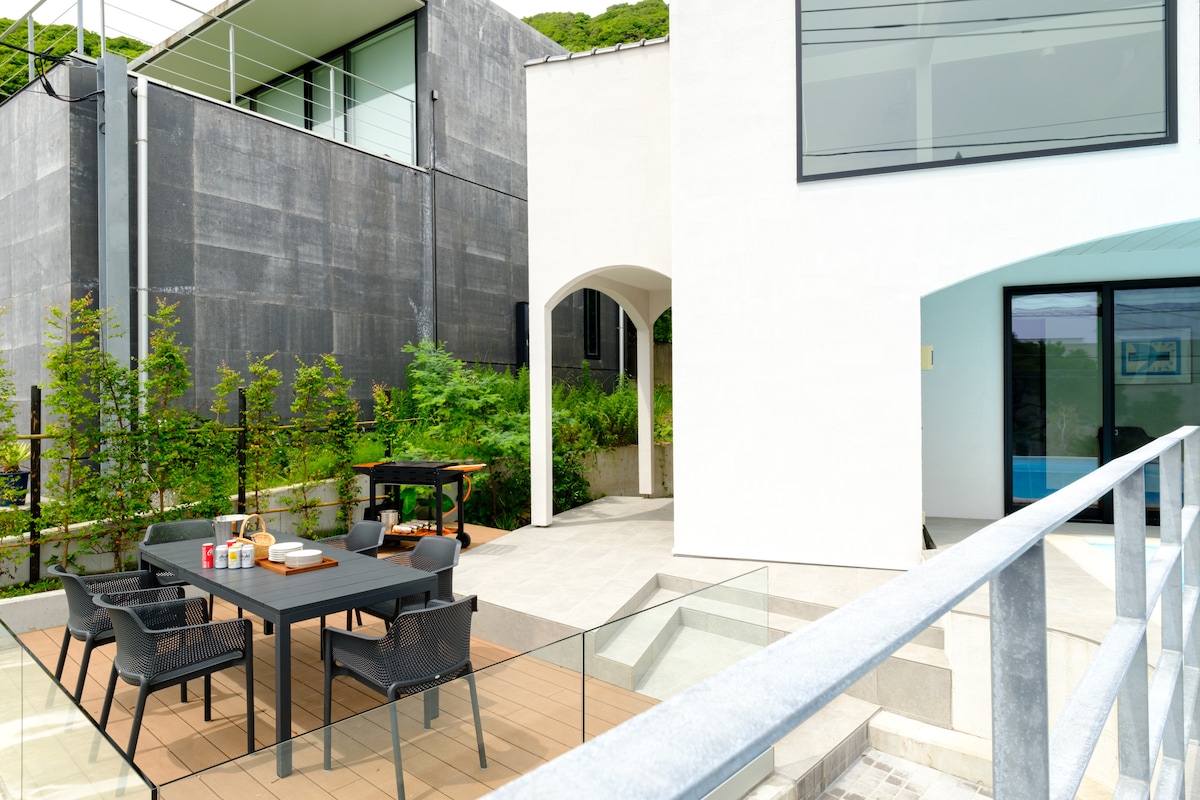 An outdoor terrace features a rectangular dining table surrounded by comfortable chairs. Lush greenery borders the space, providing a sense of privacy. A barbecue grill is visible in the background, set against modern architectural lines of the adjacent building.