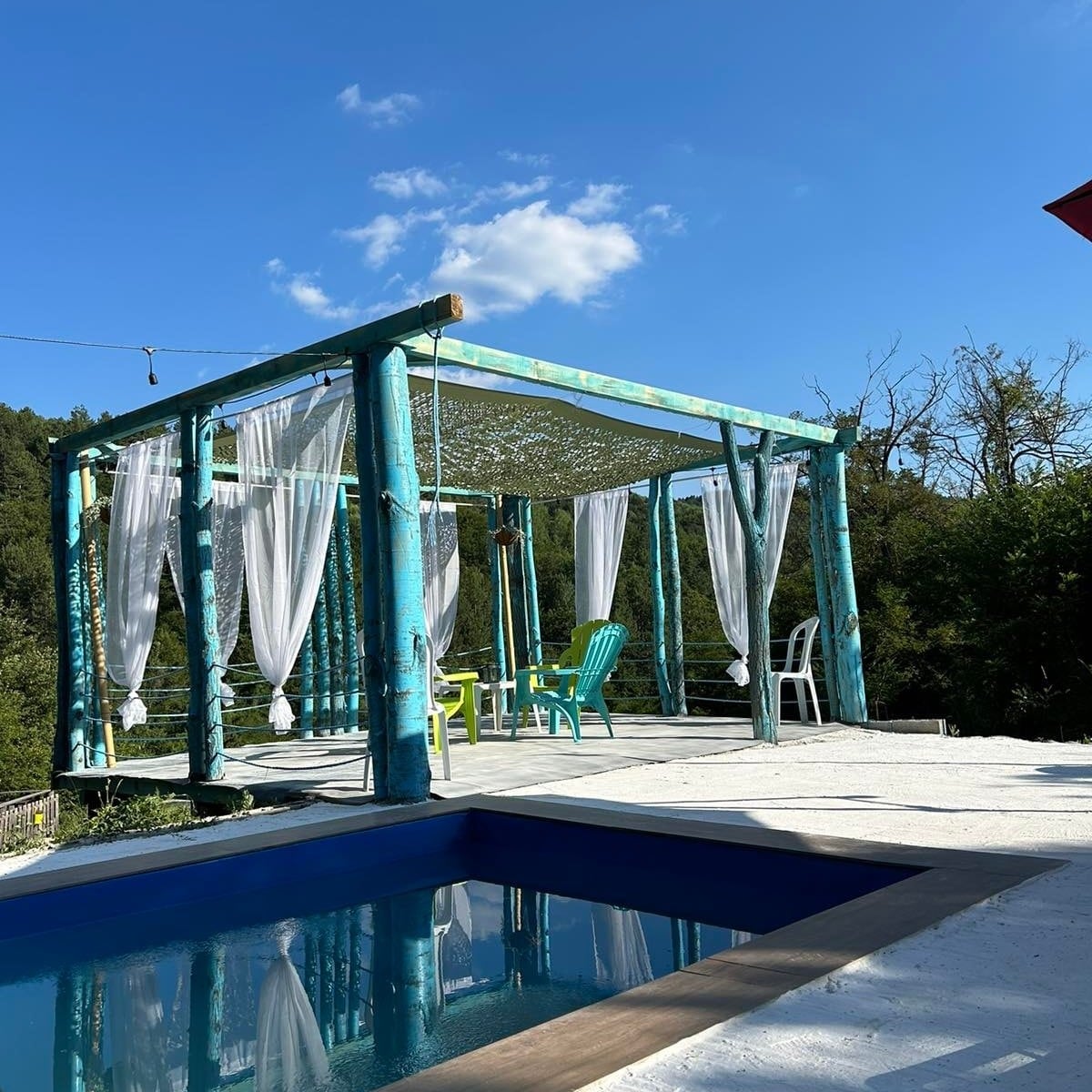 An outdoor seating area is showcased with a wooden pergola featuring flowing white curtains. Colorful chairs are positioned around a table, overlooking a tranquil blue pool. A clear blue sky and green trees complete the serene setting, inviting relaxation and leisure.
