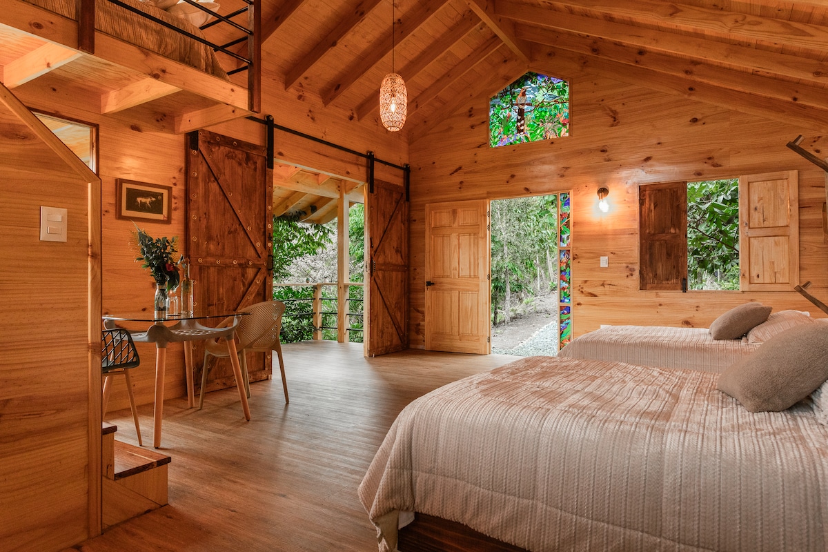 The inviting interior of the wooden cabin features two spacious beds, a dining area with chairs, and large windows showcasing natural surroundings. A warm ambiance is created by the wooden accents and warm lighting fixtures. A door leads to an outdoor area, enhancing connectivity with nature.