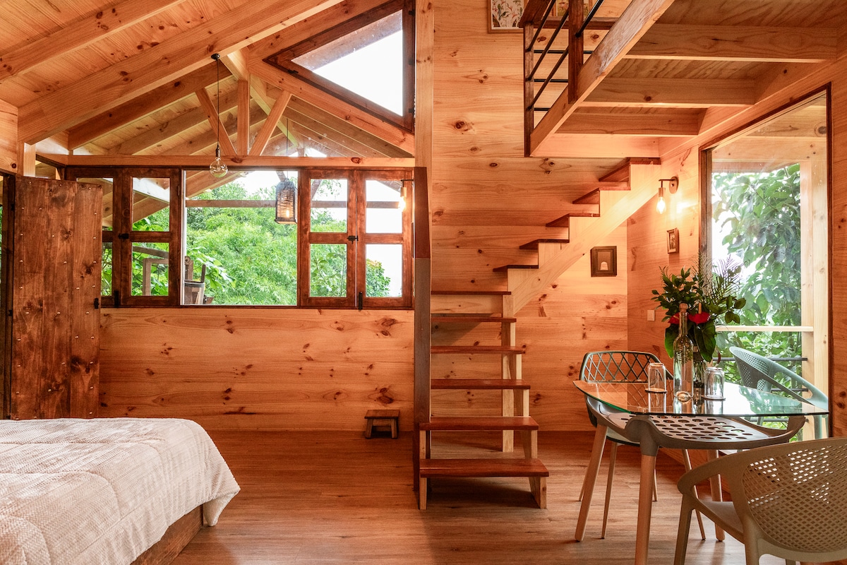 The interior showcases warm wooden walls and a staircase leading to the upper level. A spacious sleeping area features a neatly made bed with a textured blanket. A dining table is positioned near large windows, inviting natural light and views of the surrounding greenery.