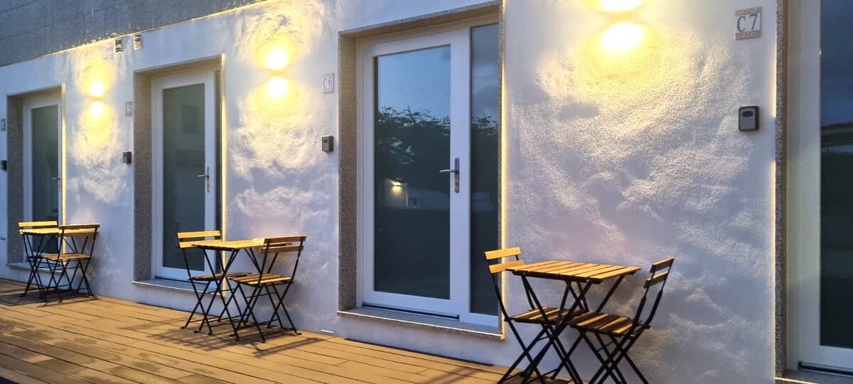 A series of private entrances is visible, featuring modern glass doors framed by smooth white walls. Two small wooden tables with matching chairs are placed on a wooden deck, illuminated by warm wall-mounted lights, creating an inviting outdoor space.