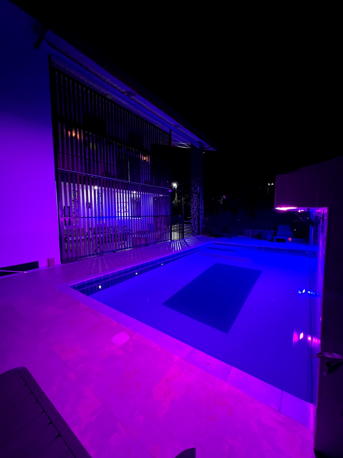 A serene pool area is illuminated by soft purple lighting, creating a calming atmosphere at night. The sleek, modern design of the surrounding architecture and spacious layout enhances the tranquil environment. Reflections of light dance on the water's surface, inviting relaxation.